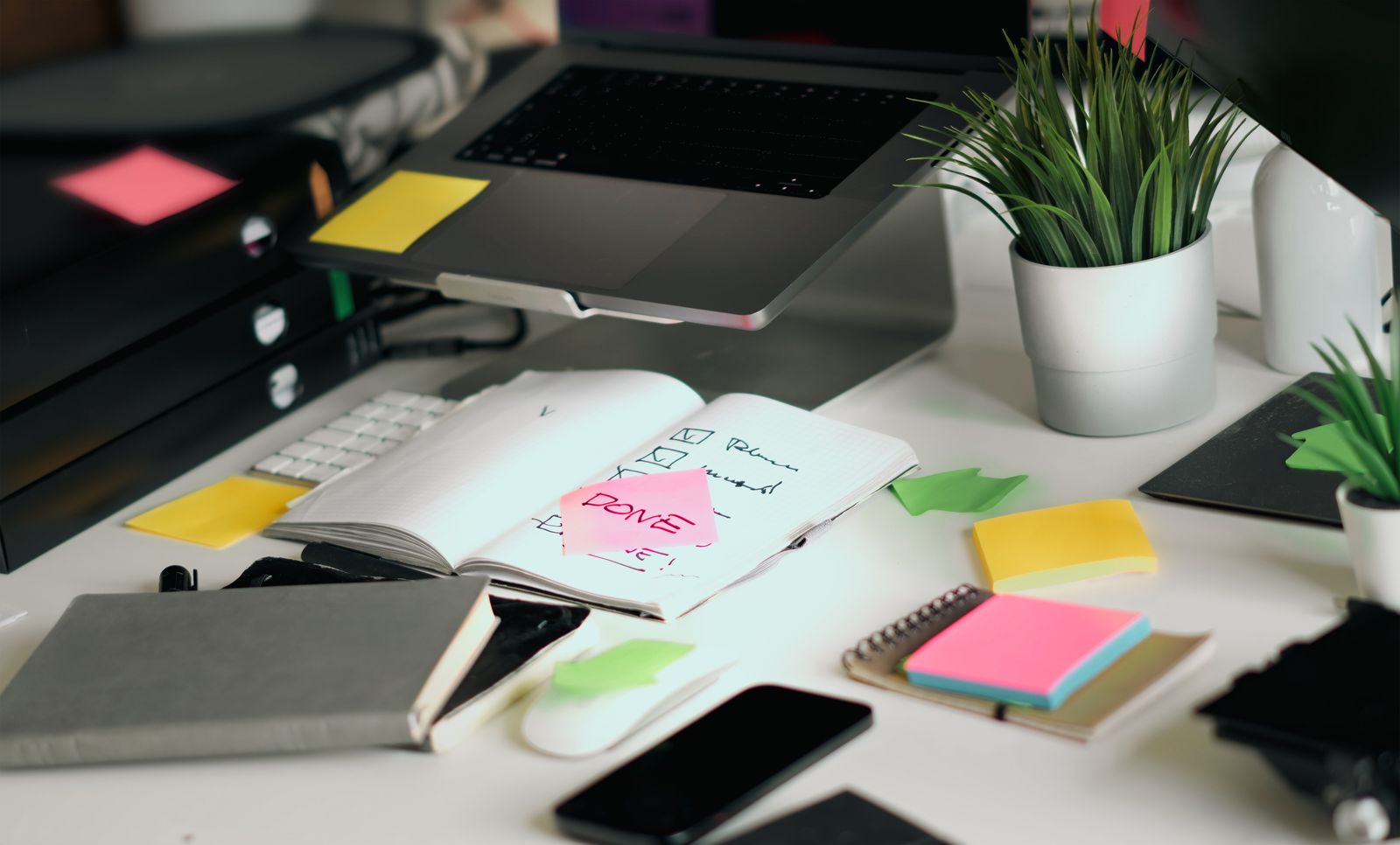 A tidy white desk with a laptop, an open notebook with a handwritten checklist and a pink sticky note reading DONE, colorful sticky notes, a phone and two small potted plants